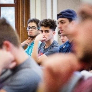 Group of people seated in a classroom or lecture hall, looking forward attentively; windows and interior details visible in the background.