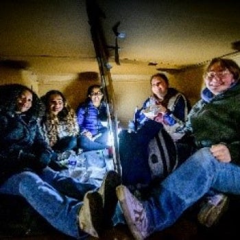 Five people sitting closely together in a dimly lit space with cardboard walls and ceiling, illuminated by a central light source; all dressed warmly.