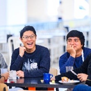 Group of people sitting around a table indoors, sharing a meal and conversing; plates and cups visible on the table.