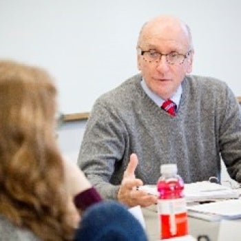 Person wearing glasses and a gray sweater with a red tie sitting at a table with papers and books, conversing with two partially visible individuals; pink bottle on the table and whiteboard in the background