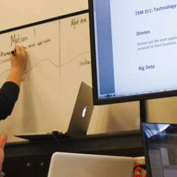 A group of students work together in a classroom setting, using a whiteboard and large display monitor