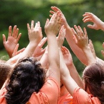 Several people wearing orange shirts standing outdoors with arms raised together in a celebratory gesture; green foliage in the background.